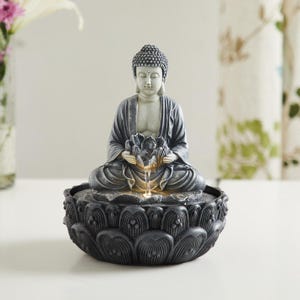 May include: A gray and black Buddha statue fountain. The Buddha is seated in a meditative pose, holding a lotus flower. Water flows from the flower into a lotus-shaped base. The statue has a serene expression and is a decorative item.