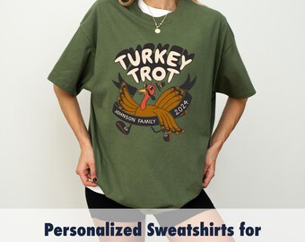 May include: An olive green t-shirt featuring a cartoon turkey design. The design includes the words "TURKEY TROT" above a turkey wearing trainers, with "JOHNSON FAMILY 2024" on a banner. The shirt is part of a family-themed collection.
