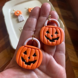 May include: A pair of orange pumpkin-shaped earrings with white handles and black carved facial features. The earrings are crafted from a smooth material and feature gold-toned metal hooks for attachment, designed to resemble miniature trick-or-treat buckets.