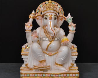 May include: A white marble statue of the Hindu deity Ganesha, seated in a regal pose. The statue features intricate gold detailing on the crown, jewelry, and base. The deity is holding a lotus flower and a sweet treat. The statue is on a decorative base.