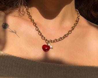 May include: Gold-toned chain necklace with a red cherry pendant and two small white pearls. The necklace has a chunky link design and is worn around the neck. The cherry pendant is glossy and vibrant.