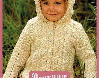 Könnte beinhalten: Ein weißer gestrickter Kapuzenpullover mit Knöpfen. Der Pullover hat ein Zopfmuster und wird von einem Kind auf einer Wiese getragen.