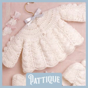 Könnte beinhalten: Ein weißer gehäkelter Baby-Pullover mit Knopfleiste und einem gewellten Saum. Der Pullover liegt auf einem rosa-weiß gestreiften Hintergrund.