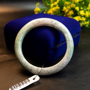 May include: A jade bangle bracelet with a pale green and white marbled appearance. The bracelet is circular and sits on a dark blue velvet surface. A white tag with a barcode hangs from the bracelet.