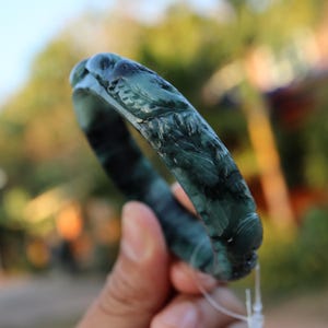 May include: A dark green jade bangle bracelet with carved floral and leaf designs. The bracelet is held up against a blurred background. The jade has a polished, glossy finish and shows variations in color.