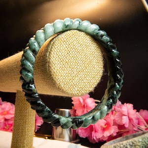 May include: A jade bracelet with a twisted rope design. The bracelet is a gradient of dark green to light green. The bracelet is displayed on a tan jewelry stand with pink flowers in the background.