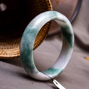 May include: A smooth, polished bangle bracelet crafted from translucent stone, featuring a rounded profile with a variegated pattern of milky white and deep forest green swirls throughout the circular band, accompanied by a small white paper tag.