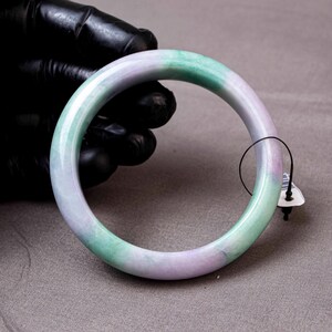 May include: A smooth, circular bangle bracelet crafted from polished stone, featuring a marbled pattern of soft lavender and light green hues. The bangle has a rounded profile and includes a small black string tag attached to one side.