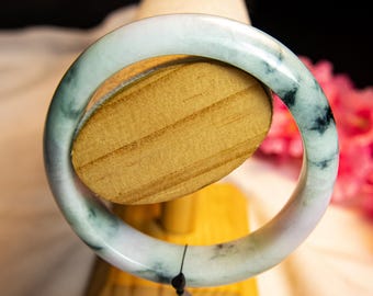 Pode incluir: Uma pulseira de jade com aparência marmorizada, apresentando tons de verde, branco e preto. A pulseira é exibida em um suporte de madeira, destacando sua superfície lisa e polida. O design circular da pulseira é realçado.