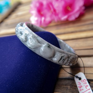 May include: A gray jade bangle bracelet with carved floral and geometric designs. The bracelet is resting on a blue velvet surface. The bracelet has a diameter of 2.2 inches.