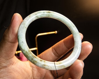Könnte beinhalten: Ein Jade-Armreif mit einer Mischung aus weißen, grünen und braunen Farbtönen. Der glatte, kreisförmige Armreif wird in einer Hand vor dunklem Hintergrund gehalten. Der Armreif hat einen Durchmesser von etwa 6,35 cm.
