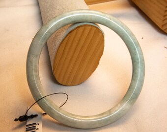 Könnte beinhalten: Ein hellgrünes Jade-Armreif-Armband. Das glatte, kreisförmige Armband wird auf einem Schmuckständer aus Holz präsentiert. Das Armband hat eine subtile, natürliche Farbvariation und eine polierte Oberfläche. Das am Armband befestigte Etikett lautet AU092.