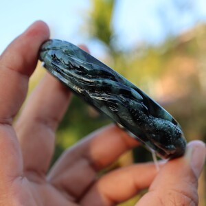 May include: Dark green jade bangle bracelet with intricate dragon carving. The bracelet is held in a hand, showcasing the detailed relief work and the polished surface of the stone. The dragon design is visible along the length of the bracelet.