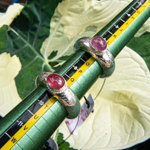 May include: Two silver rings with oval, red gemstones are displayed on a green ring sizer. The rings have ornate silver detailing around the gemstones. The ring sizer has black and yellow markings for ring sizes.