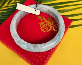 Könnte beinhalten: Ein hellgrünes und weißes Jade-Armreif-Armband liegt auf einer roten Schachtel mit einem goldenen Blumendesign. Das Armband ist rund und glatt, mit marmoriertem Aussehen. Ein leeres Etikett hängt am Armband.