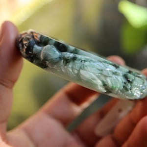 May include: A carved jade bracelet in shades of green and black. The bracelet features a detailed relief carving of a figure. The jade is polished and has a translucent quality, with variations in color and texture.