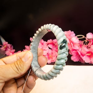 May include: A light green jade bracelet with a twisted rope design. The bracelet has a smooth, polished finish and a touch of dark green veining. The background features pink flowers.