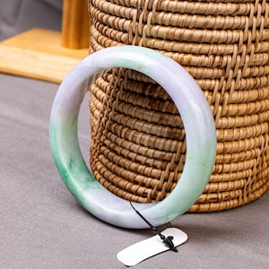 May include: A smooth, circular bangle bracelet crafted from polished stone, featuring a translucent appearance with blended swirls of pale lavender and vibrant green hues. A small white rectangular tag is attached by a thin black plastic cord.