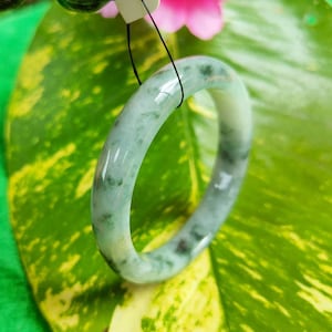 Può includere: Un braccialetto a cerchio di giada con una superficie liscia e lucida. Il braccialetto è di colore verde chiaro con inclusioni verdi e marroni più scure. Il braccialetto è esposto su una foglia verde e gialla.
