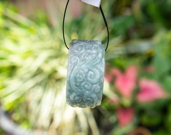 May include: A light green jade pendant with intricate swirling patterns carved into its surface. The cylindrical pendant is suspended from a black cord, showcasing the detailed craftsmanship. The background is a soft blur of green foliage.