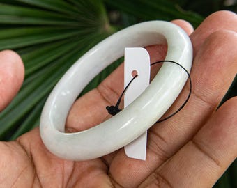 Könnte beinhalten: Ein hellgrünes Jade-Armreif-Armband. Das glatte, abgerundete Armband wird in einer Hand gehalten, mit einem weißen Anhänger. Der Hintergrund zeigt grünes Laub.