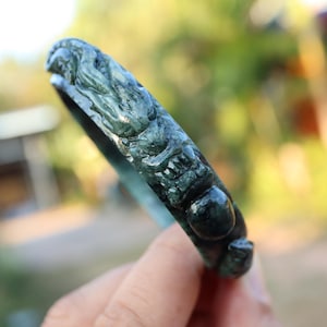 May include: A dark green jade bracelet with intricate carvings of mythical creatures. The bracelet has a smooth, polished finish and a rounded shape. The carvings are detailed and add a unique touch to the jewelry.
