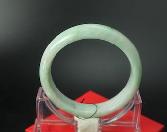 Könnte beinhalten: Ein hellgrünes Jade-Armreif-Armband. Das kreisförmige Armband wird auf einem transparenten Acrylständer mit roter Basis präsentiert. Der Jade hat eine transluzente Qualität mit Farbvariationen. Der Durchmesser des Armbands beträgt ca. 6,5 cm.