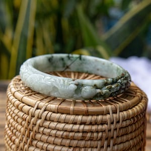 May include: A jade bangle bracelet with carved floral and dragon motifs. The bracelet is primarily light green with darker green and black accents. The bracelet is resting on a woven basket.