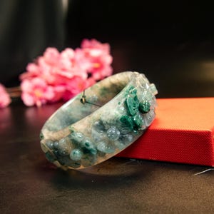 May include: A jade bangle bracelet with carved floral designs. The bracelet features a mix of green and white hues, with detailed flower carvings around the band. The bracelet is displayed on a red surface with pink flowers in the background.