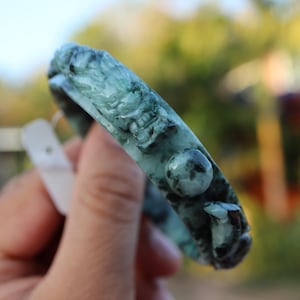 May include: A carved jade bracelet in shades of green and black. The bracelet features detailed relief carvings of a dragon and other elements. The jade has a polished finish and is held by a hand.
