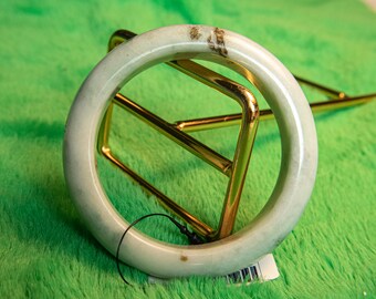 Könnte beinhalten: Ein hellgrünes Jade-Armreif-Armband mit braunen Markierungen. Das Armband wird auf einem goldfarbenen Metallständer vor einem hellgrünen Hintergrund präsentiert. Der Jade hat eine glatte, polierte Oberfläche.