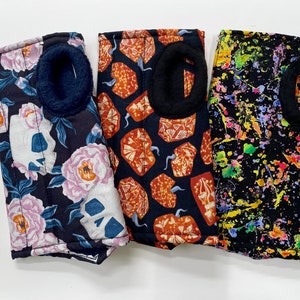 May include: Three fabric face masks with different patterns. The first mask has a floral and skull design. The second mask has a geometric pattern with orange and brown shapes. The third mask has a black background with colourful paint splatters.