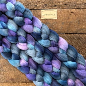 May include: A braided skein of Corriedale wool in shades of blue, purple, and lavender. The wool is arranged on a wooden surface, with a small label that reads "Corriedale". The colors blend together, creating a soft, textured appearance.