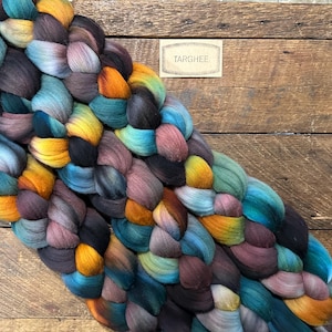 May include: A colorful braid of Targhee wool roving, featuring a mix of blues, greens, yellows, oranges, and purples. The wool is arranged in a thick, textured pattern. A small label with the word "Targhee" is visible.
