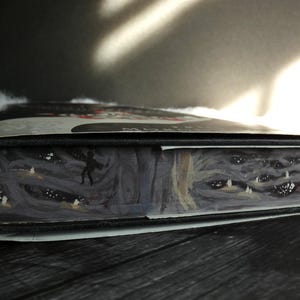 May include: Close-up of the spine of a hardback book with a black cover and a white and grey illustration of a forest scene with a figure walking through it. The illustration also features a close-up of a creature's face with multiple eyes.