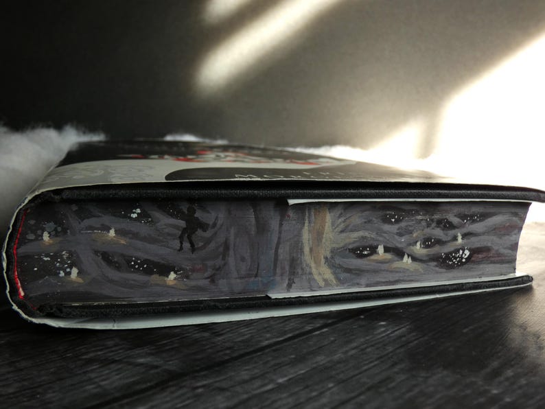 May include: Close-up of the spine of a hardback book with a black cover and a white and grey illustration of a forest scene with a figure walking through it. The illustration also features a close-up of a creature's face with multiple eyes.