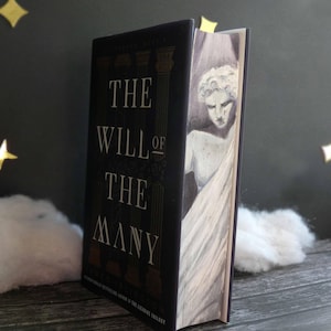 Op de afbeelding: Een hardcover boek getiteld "The Will of the Many" met gouden accenten en een donkere omslag. De rand van het boek heeft een handgeschilderde illustratie van een klassiek standbeeld. De achtergrond is donker met stervormige decoraties.