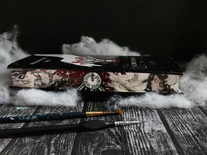 A hardcover book of Erin Morgenstern's The Night Circus with the edge of the pages painted, depicting two portraits facing eachother, the left with branches behind him, the right with birds behind her. A clock sits in the middle.