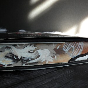 May include: Close-up of a hardback book with a black cover and a painted illustration of a white creature with a black eye on the front. The illustration is on a brown background with white stripes.