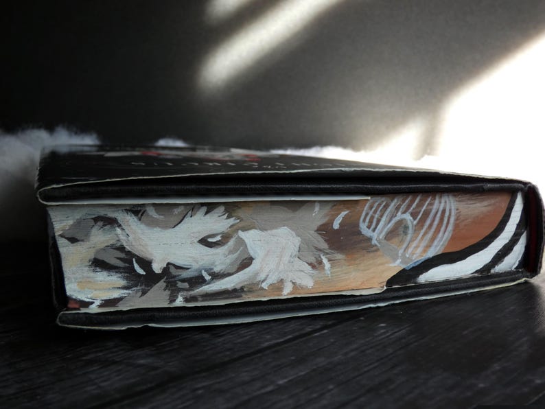 May include: Close-up of a hardback book with a black cover and a painted illustration of a white creature with a black eye on the front. The illustration is on a brown background with white stripes.