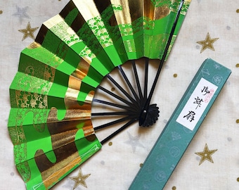 May include: A folding hand fan with alternating panels of bright green and metallic gold, featuring black line-art patterns of pine trees. The fan has black ribs and is displayed next to a rectangular green box with white text reading 扇子.