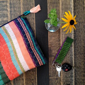 Peut inclure: Une pochette tiss&eacute;e color&eacute;e avec un pompon p&ecirc;che, une dragonne verte et violette avec des cl&eacute;s, un petit vase en verre avec de la menthe et une fleur jaune &agrave; centre noir, sur une surface en bois.
