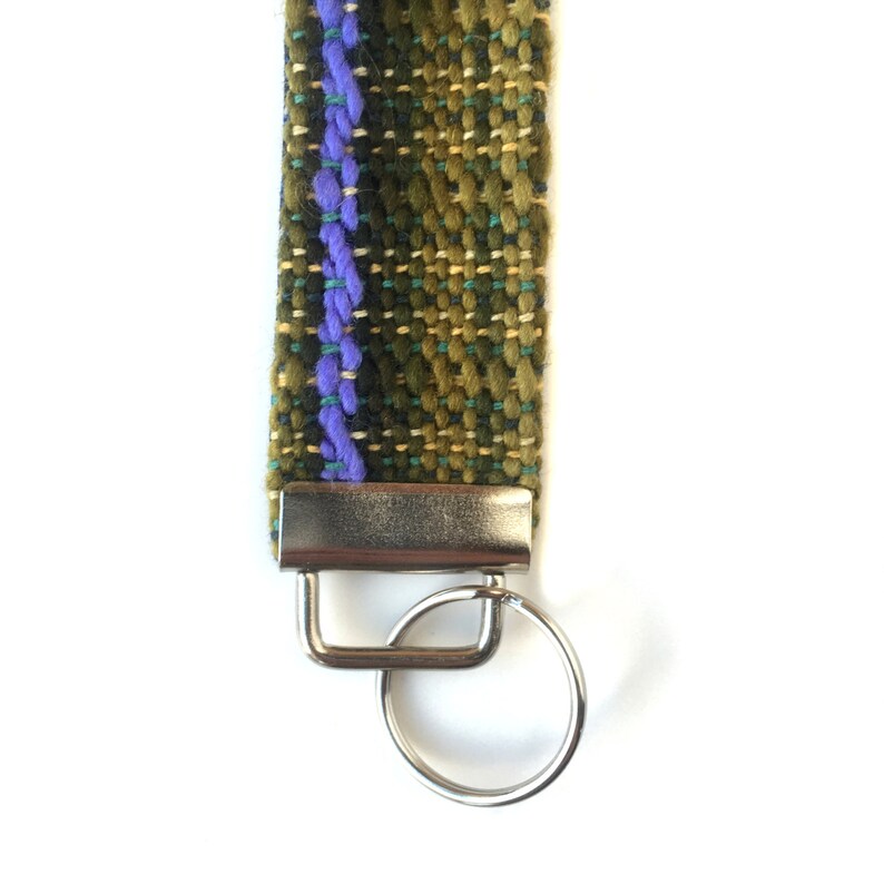 May include: A woven key fob with a green, olive, and blue pattern, accented by a central purple stripe. The fob is attached to a silver-colored metal clasp and key ring, set against a white background.