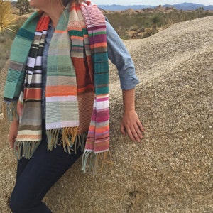 May include: A colorful, handwoven scarf with a striped pattern. The scarf features a variety of colors, including teal, orange, pink, and brown, with fringed edges. The scarf is draped over a person's shoulders.