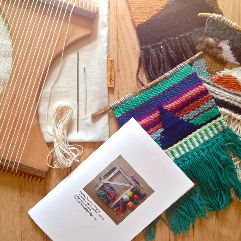 May include: A collection of weaving supplies and finished weavings are arranged on a wooden surface. The items include a wooden loom, needles, yarn, a canvas bag, and a booklet. Several colorful weavings with fringe are also present.