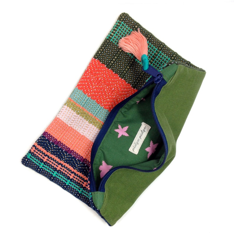 Peut inclure: Une pochette tiss&eacute;e color&eacute;e avec un ext&eacute;rieur vert et une fermeture &eacute;clair bleue. La pochette pr&eacute;sente un motif ray&eacute; dans les tons orange, rose, vert et bleu. L'int&eacute;rieur est vert avec des motifs d'&eacute;toiles roses et une &eacute;tiquette "pildge pildge". Un pompon rose pend de la fermeture &eacute;clair.