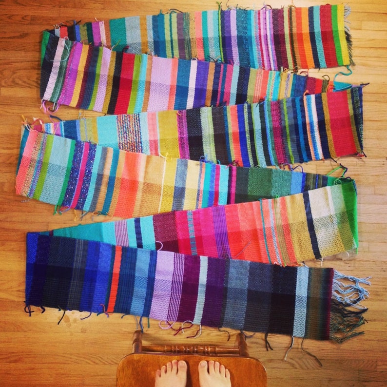 May include: Five colorful, handwoven scarves with vertical stripes in various colors, including blue, pink, green, and yellow. The scarves are laid out on a wooden floor, showcasing their vibrant patterns and textures. Each scarf has fringe at the ends.