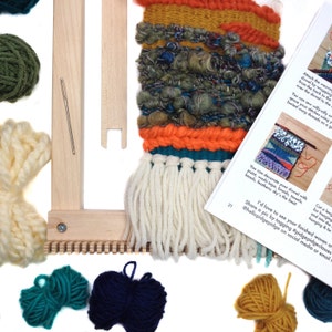 May include: A colorful weaving project in progress, featuring a wooden loom, yarn in various colors, and a partially completed woven piece. The woven art includes orange, teal, and cream-colored fringe. A booklet with instructions is also visible.