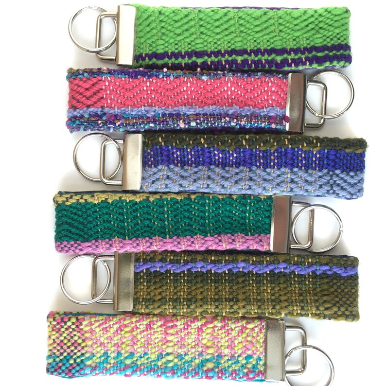 May include: A collection of six woven key fobs in various colors. The key fobs feature a rectangular shape with a silver-colored metal clasp and key ring. The color palette includes green, pink, blue, and olive green, with purple and yellow accents.