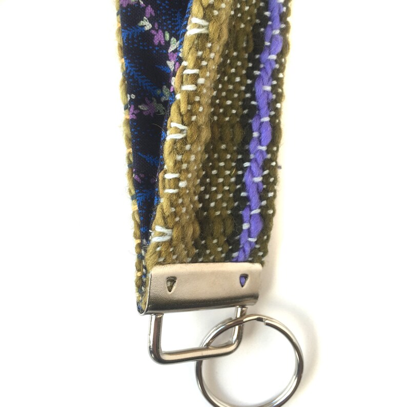 May include: A handmade wrist lanyard featuring a mix of patterned fabrics in blue floral, olive green, and purple. The lanyard is attached to a silver-colored metal clasp and key ring. The design is a mix of woven and stitched textures.